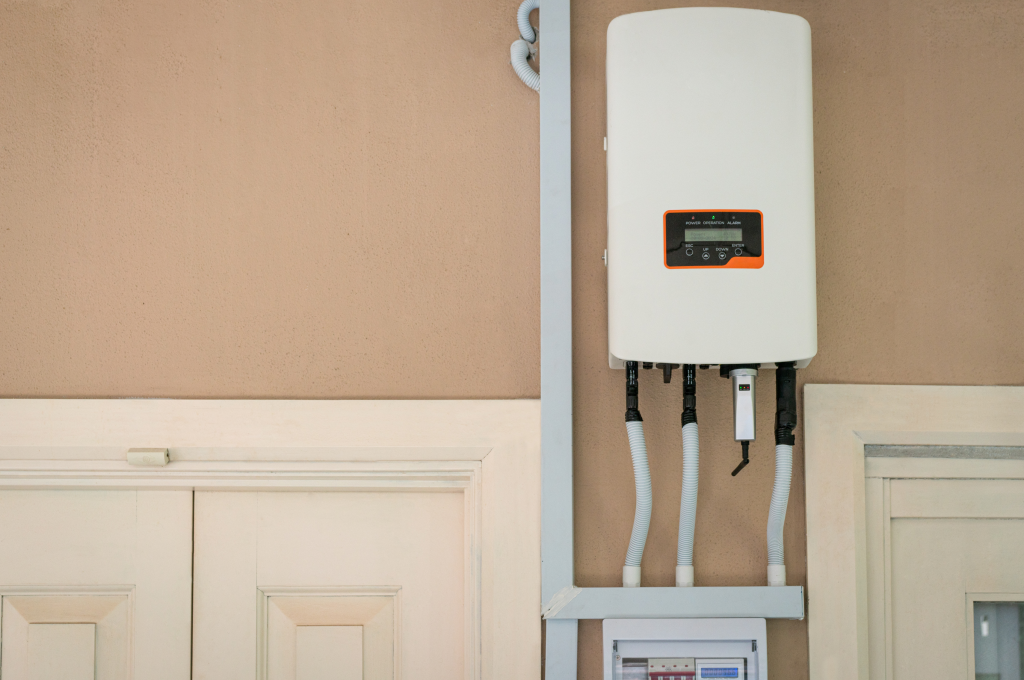 - Inverter rettangolare montato su muro esterno vicino a una porta, con cavi elettrici collegati. Questo dispositivo trasforma la corrente continua dei pannelli in corrente alternata utilizzabile in casa.